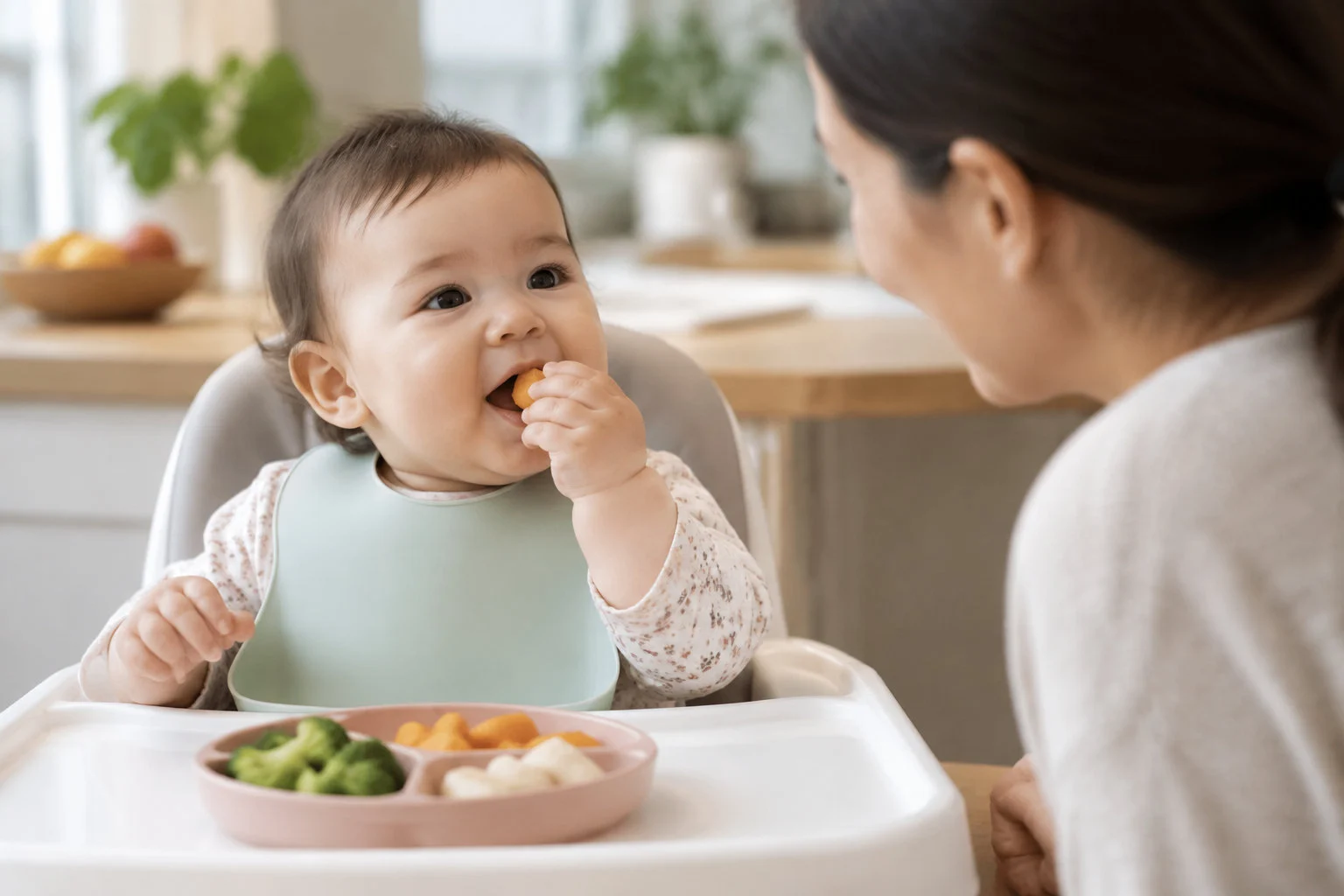 Un bébé assis dans une chaise haute porte un aliment à sa bouche, tandis qu’une femme le regarde ; une assiette compartimentée contenant des légumes et des morceaux de nourriture est posée devant lui.